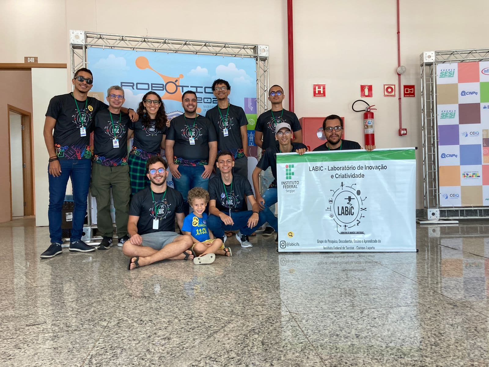 Equipe do LABIC reunida no palco da feira Robótica 2023 com o banner do laboratório, no Instituto Federal de Sergipe